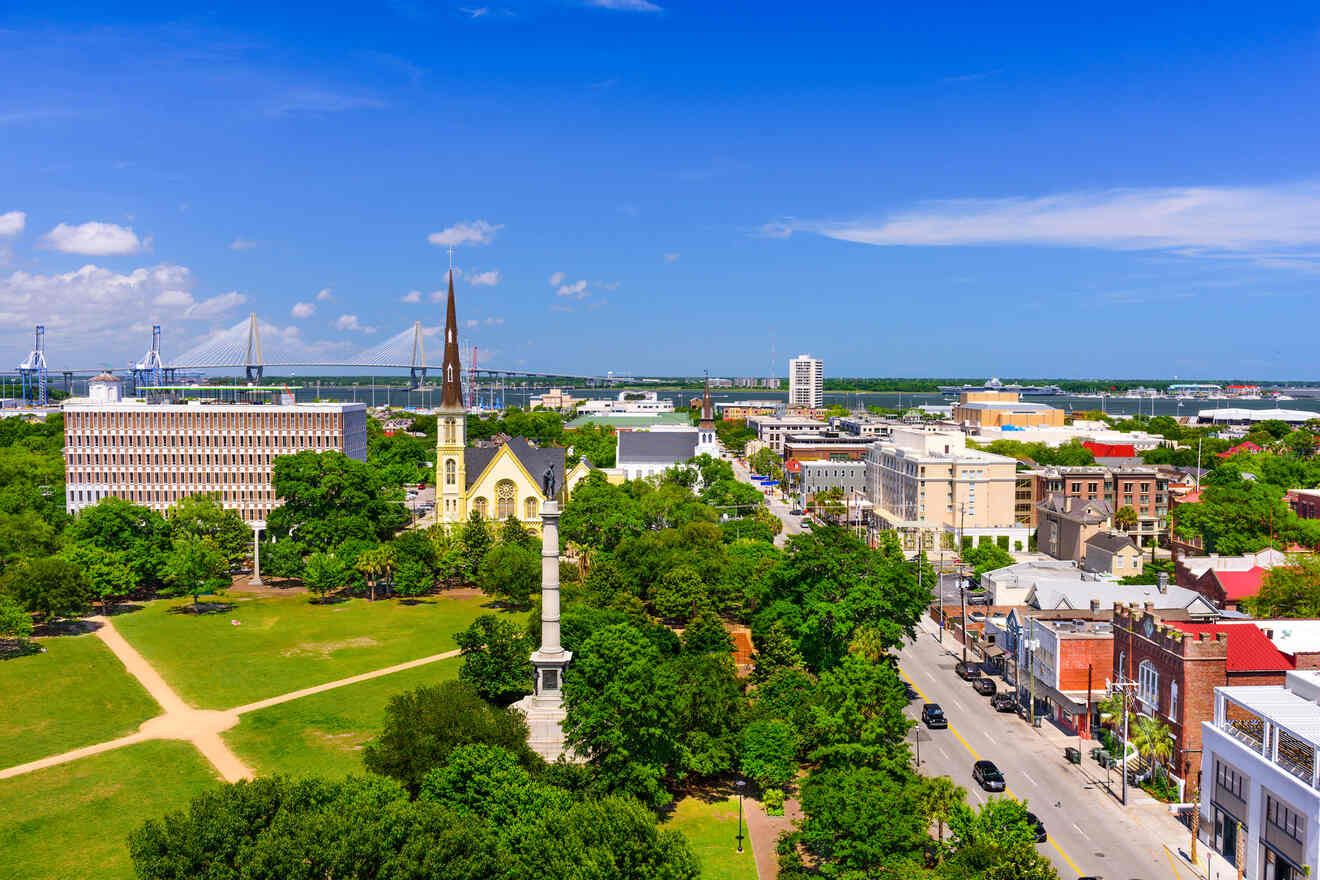 Aerial view of Charleston, South Carolina, showcasing the Ravenel Bridge in the distance, historic church spires, and the lush Marion Square Park leading to a bustling urban street.